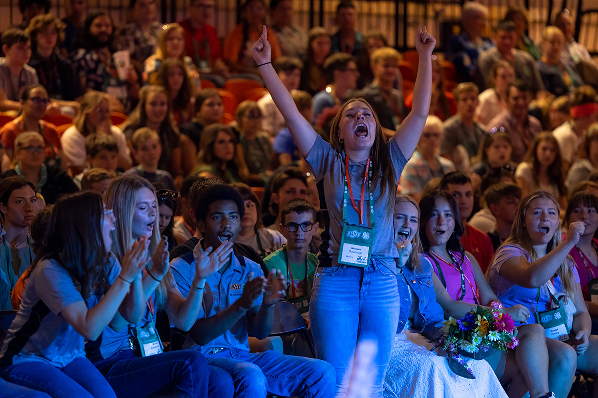 A 4-H member stands up, arms raised in celebration, in front of a crowd at Roundup.
