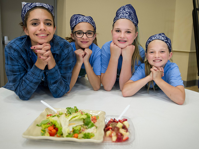 4-H members lean forward smiling while helping serve others food. Girls showing their food in food showdown